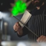 Close-up of a chef sharpening a kitchen knife with a honing steel in a professional kitchen setting.